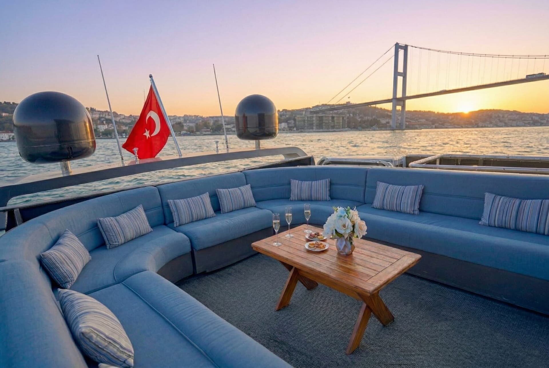 Blue lounge sofas on the open deck of a Luma charter yacht, the Bosphorus Bridge in golden evening light beyond